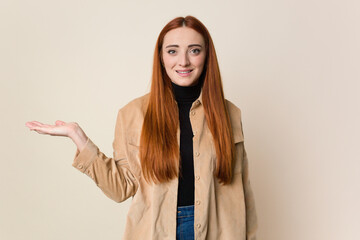 Young red hair woman isolated showing a copy space on a palm and holding another hand on waist.