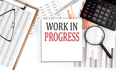 WORK IN PROGRESS text on notebook with clipboard and calculator on a chart background