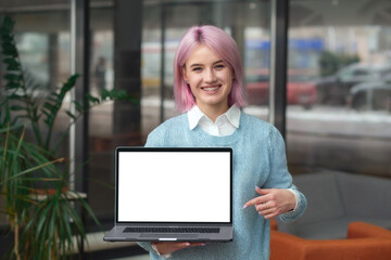 Happy successful young woman student, programmer or entrepreneur holds laptop in his hands, points his finger at blank screen stands in modern office or coworking, looking at the camera and smiling