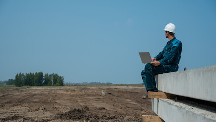 Caucasian male builder in hardhat sits on floor slabs and uses laptop at construction site. 