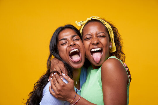 Two diversity pretty positive girls stick out your tongue hugging isolated on yellow color background. Young couple goofing around embracing and looking camera. Friendship and homosexual relationship.