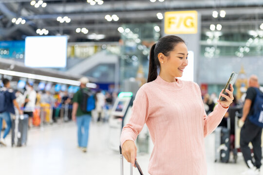 Asian woman holding passport and luggage with using mobile phone walking to airline check in counter in airport terminal. People travel on holiday vacation and global airplane transportation concept.
