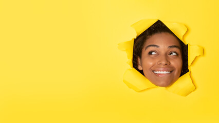 Happy black lady posing in hole in torn paper looking aside, advertising great offer over yellow background, copy space