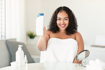 Plus size woman showing shampoo bottle, advertising beauty product and smiling, sitting wrapped in towel after bath