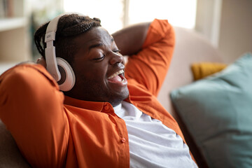 Joyful black guy reclining on sofa, singing songs, using headphones