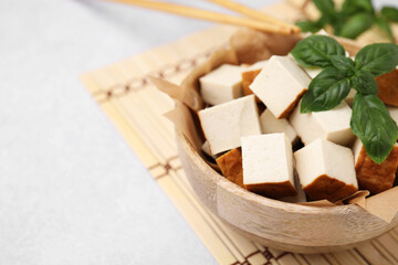 Bowl of smoked tofu cubes and basil on white table, closeup. Space for text