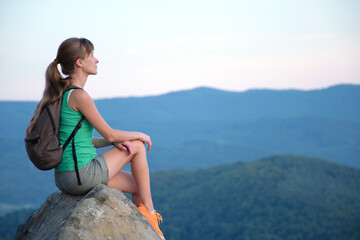 Naklejka na ściany i meble Sportswoman sitting alone taking a break on hillside trail. Female hiker enjoying view of evening nature from rocky cliff on wilderness path. Active lifestyle concept
