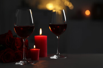 Glasses of red wine, burning candles and rose flowers on grey table against blurred lights, space for text. Romantic atmosphere