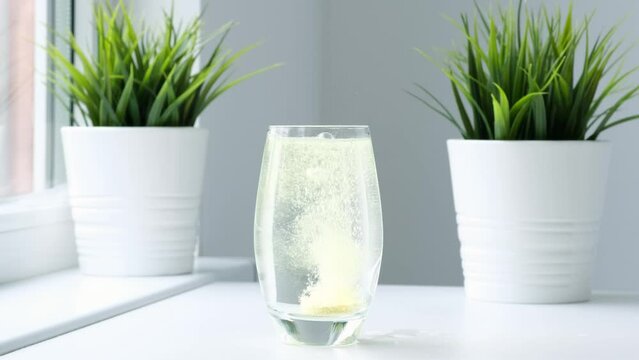 Effervescent pill dissolves with bubbles in water on white table. Vitamins C pills against flu, medicines, treatment at home. Vitamin tablet falling down in glass and colors water yellow. Slow motion