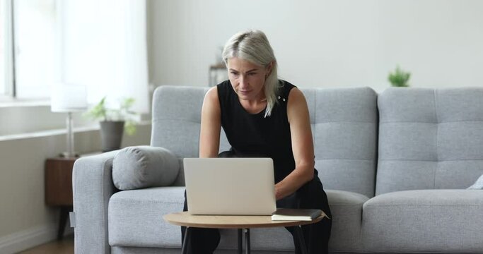 Thoughtful mature woman typing on laptop keyboard, sits on sofa consider answer to client, lead correspondence, working from home use computer, search solution, looks focused while do telework on-line