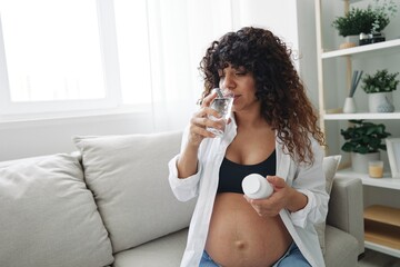 Fototapeta na wymiar Pregnant woman smile and happiness taking pills and vitamins for pregnant women with pills hand water drinks clean water, maintaining water balance in the body, swelling, sitting on the couch at home