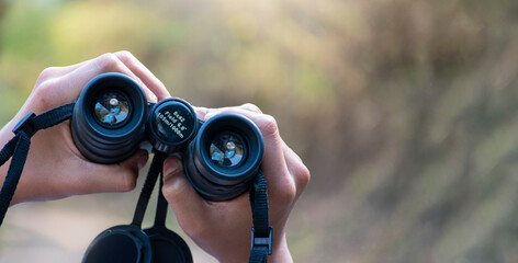 Closeup view of binoculars holding in hands of young asian natural surveyor, blurred dry autumn national forest park background.