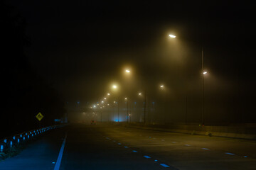 View dark night foggy misty highway city road and street lights low poor visibility cold spring autumn season. Seasonal bad weather accident danger warning car fog light.