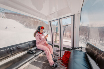 Woman skier using phone app in gondola ski lift on ski holidays. Girl smiling using mobile smartphone wearing cool ski clothing, helmet and goggles. Ski winter vacation activity concept © Maridav