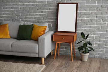 Wooden table with blank frame, houseplant and sofa near grey brick wall