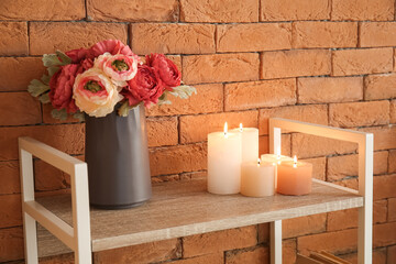 Burning candles and vase with beautiful flowers on shelving unit near brick wall