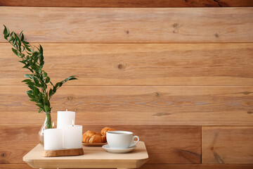 Burning candles, cup of coffee, croissants and vase with ruscus branches on end table near wooden wall