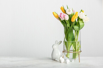 Glass vase with beautiful tulip flowers, Easter eggs and ceramic bunny on light background