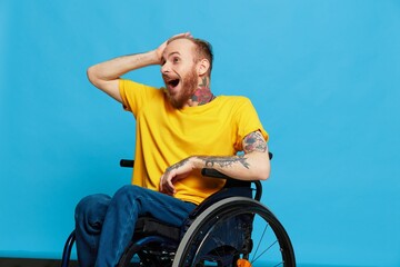 A man in a wheelchair surprised and open mouth in a t-shirt with tattoos on his arms sits on a blue studio background, a full life, a real person