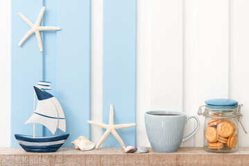 Summer sea decoration background - Front view of sailboat , starfish , seashell with coffee cup and cookie on shelf over blue white wall .