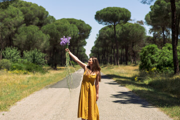 A young woman with straight black hair, a yellow summer dress standing on a road in the woods raises a bouquet of wild flowers with one hand