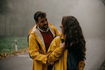 Happy affectionate couple in raincoats enjoying in nature, standing on the mountain road, and man zipping woman's backpack.