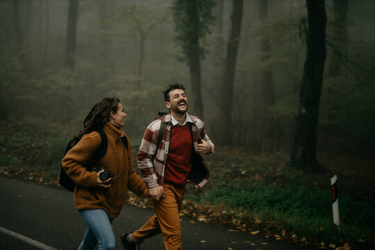 A young couple of backpackers and trekkers holding hands and running and smiling during a rainy day in the woods.