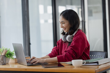 Happy smiling young asian business woman wear headphones working online sitting at desk in the office,