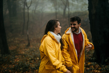 Fototapeta premium Affectioned couple in raincoats holding hands and having fun during a foggy day in the forest.