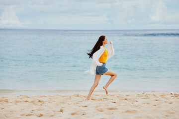 Sports woman runs along the beach in summer clothes on the sand in a yellow T-shirt and denim shorts white shirt flying hair ocean view