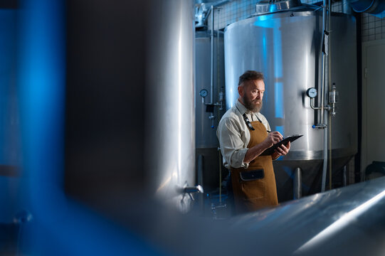 Handsome bearded man brewer inside modern beer factory around steel tanks