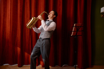 Young attractive musician plays tenor saxophone on stage with red drape curtains