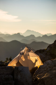 Man in tent reaches hand out, shadow of fingers toward mountain peaks.