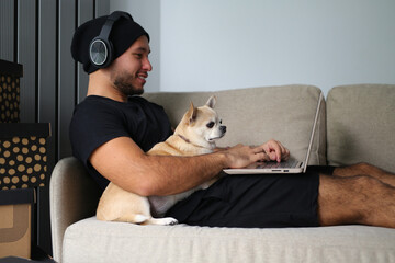 A stylish guy dressed in black clothes, with headphones, at home with his dog, work on a laptop,...