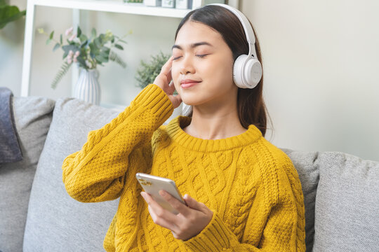 Relaxing Asian Young Woman Using Smart Phone Listening To Music, Enjoy Song Or Watching Videos, Podcast On Mobile With White Wireless Headphones Sitting On Sofa, Couch At Home, Chill Out And Leisure.