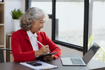 Asian senior manager working on tablet with laptop sitting at desk in office. 