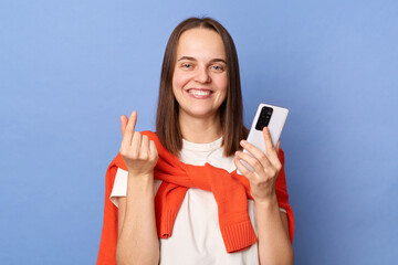Naklejka premium Image of charming smiling Caucasian woman wearing white T-shirt and orange sweater tied over shoulders standing isolated on blue background, using mobile phone, showing love gesture with fingers.