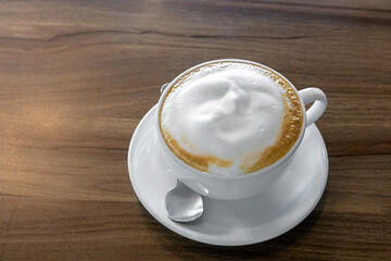 hot coffee with milk foam topping in white cup and small spoon in brown wood table