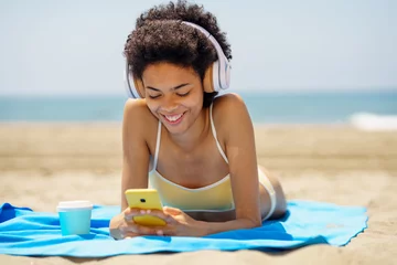 Poster Delighted ethnic female tourist in headphones browsing smartphone on seashore © javiindy