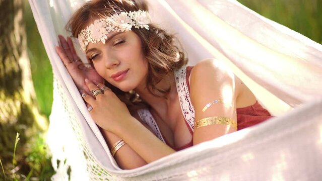 Girl in boho style clothing sleeping peacefully while lying in a hammock in a park on a sunny summer afternoon
