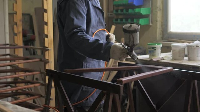 Slow motion. Carpenter-men wearing gloves, apply varnish or primer with compressed air on a wooden surface. Painting of wooden products with a spray gun in a carpentry workshop at a furniture factory