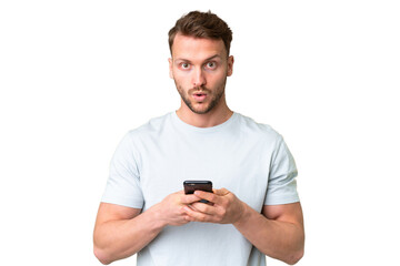 Young handsome caucasian man over isolated chroma key background looking at the camera while using the mobile with surprised expression