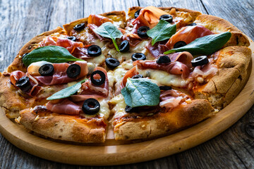 Circle prosciutto pizza with mozzarella, black olives and spinach leaves on wooden table
