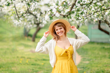 Portrait of a beautiful girl in a yellow dress in a blooming garden. A young woman in a braided hat smiles in spring