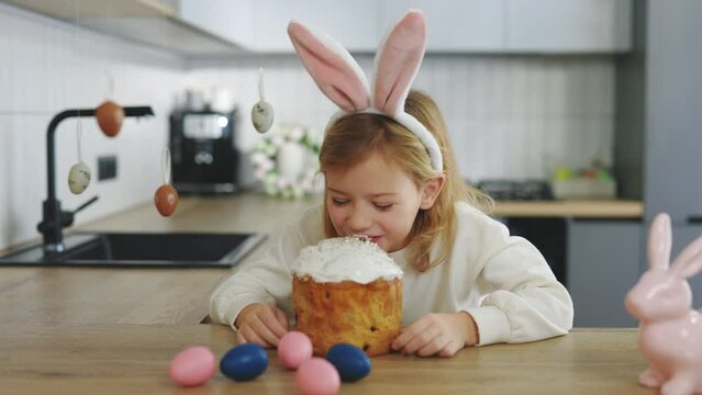 Little girl wearing rabbit ears sitting in the kitchen table and eating easter cake. Happy kid celebrating Easter with colored eggs and easter cake. Funny bunny child holds an Easter cake in her hands