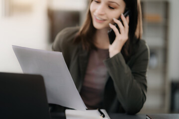 woman talking mobile phone and working sitting at her desk, reading stats and graphs on paperwork at the office.