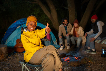 Portrait of a young black girl giving thumbs up happy with her friends in the background camping outdoors at night. Wellness and travel concept.