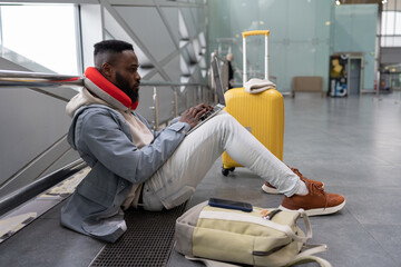 Fototapeta na wymiar Pensive African American man freelancer traveler with yellow suitcase concentrating on network, working on laptop, communicating in social media, searching information online while sitting in airport.