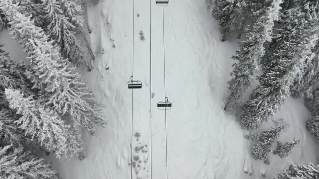 Aerial view over snowy mountain winter forest with chair lift at ski resort. Ski lift cable car top down drone shot. Winter landscape scene with snow covered trees. Outdoor tourism skiing snowboarding