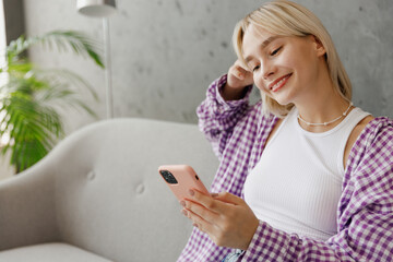 Young smiling happy woman wear casual clothes hold use mobile cell phone sits on grey sofa couch stay at home hotel flat rest relax spend free spare time in living room indoor. People lounge concept.